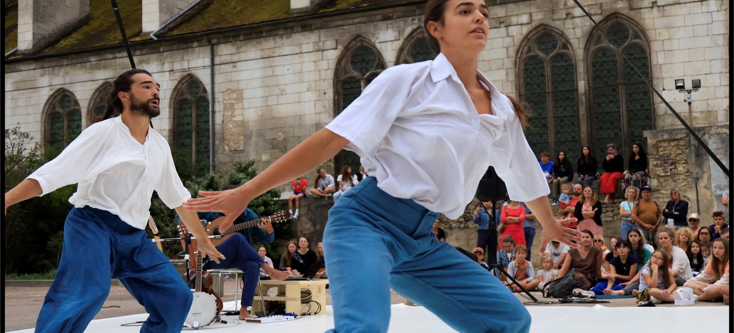 Deux acrobates en pantalon de travail bleu et chemise blanche courent l'un vers l'autre sur un tapis blanc posé devant une église, le musicien et le public assis derrière eux